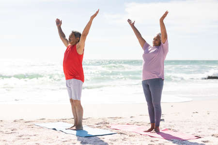 Full length of biracial senior couple practicing yoga with arms raised at sunny beach against sky. active lifestyle and fitness.の写真素材