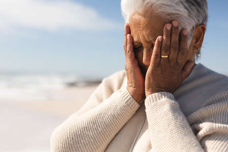 Worried retired senior biracial woman with hands covering face at beach on sunny day. lifestyle and...