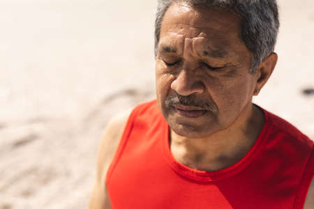 Senior biracial retired man meditating with eyes closed at beach on sunny day. active lifestyle and fitness.の写真素材