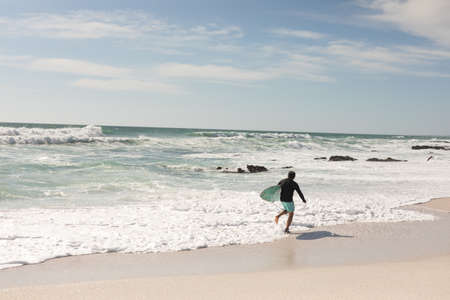 Biracial senior man with surfboard running along shore at beach on sunny day. water sport and active...