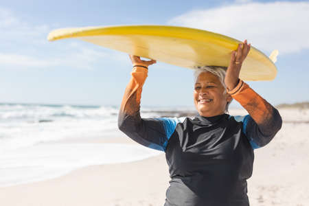 Happy biracial senior woman enjoying retirement while carrying surfboard on head at sunny beach....