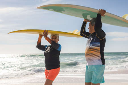 Happy multiracial senior couple looking at each other while carrying surfboards on heads at beach....