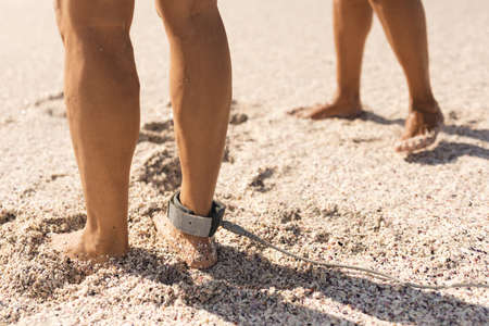 Low section of biracial senior man with surfboard leash on ankle by woman on sand at sunny beach. water sport and active lifestyle.の写真素材