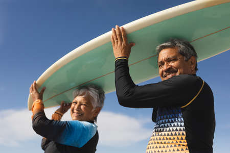 Low angle portrait of happy multiracial senior couple carrying surfboard over heads against sky. water sport and active lifestyle.の写真素材