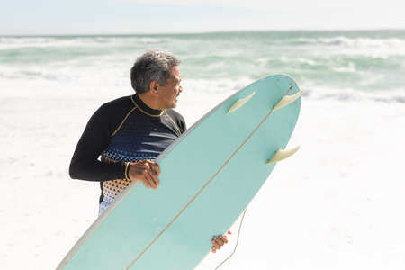 Biracial senior man carrying blue surfboard on shore while looking at sea during sunny day. water...