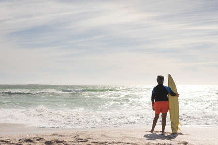 Full length rear view of senior biracial woman holding surfboard looking at horizon from sunny...