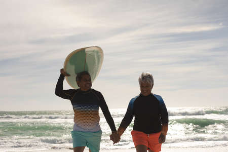 Smiling biracial senior man carrying surfboard on head while holding hand of woman walking at beach....