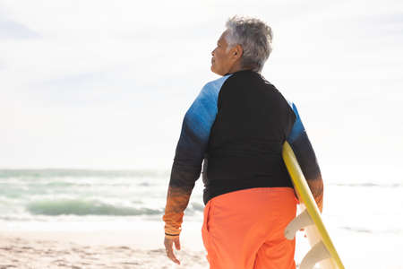 Rear view of senior biracial woman carrying surfboard looking away at beach on sunny day. water...