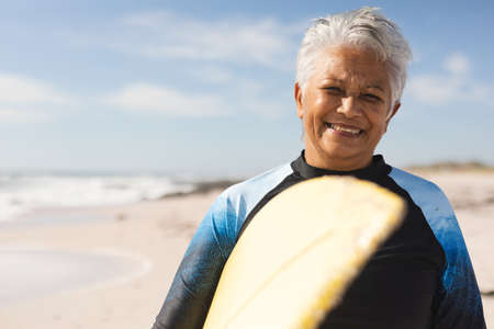Portrait of smiling biracial senior woman carrying surfboard at beach against sky on sunny day....