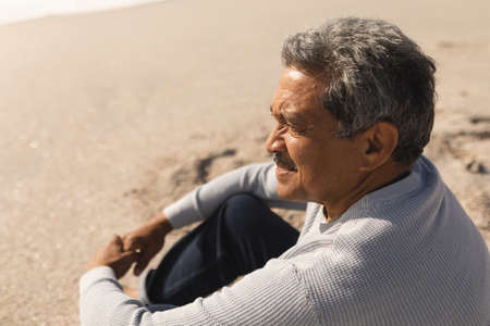Side view of retired biracial senior man looking away while sitting at beach on sunny day. active lifestyle and weekend.の写真素材