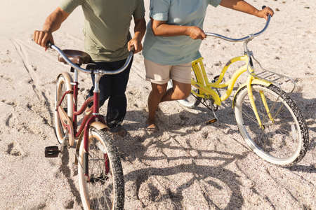 Low section of multiracial senior couple wheeling bikes on sand at beach during sunny day. active lifestyle and transportation.の写真素材