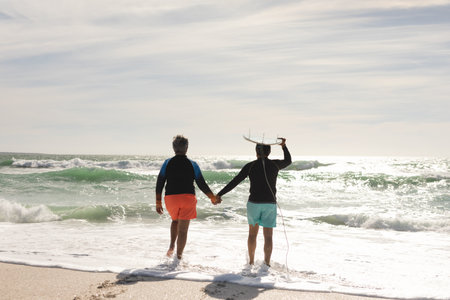 Rear view of biracial senior man carrying surfboard on head holding hand of woman walking at beach. water sport and active lifestyle.の写真素材