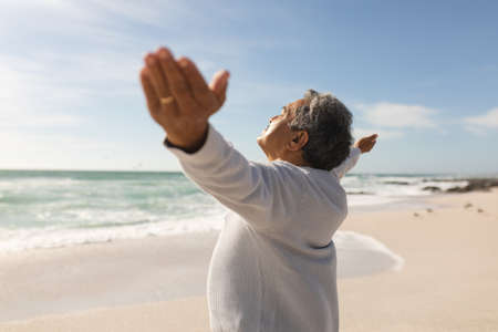 Side view of retired senior biracial man standing with arms outstretched at beach on sunny day. active lifestyle and weekend.の写真素材