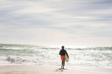 Rear view of senior biracial woman holding surfboard running towards sea at beach on sunny day. water sport and active lifestyle.の写真素材