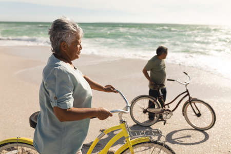 Side view of biracial senior woman and man with bicycles at beach looking away on sunny day. active lifestyle and transportation.の写真素材