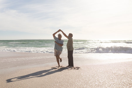 Full length of happy retired senior multiracial couple dancing on shore at beach during sunny day. lifestyle, love and weekend.の写真素材