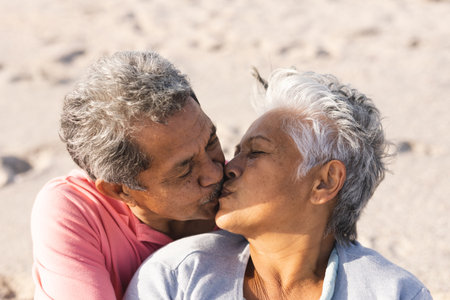 Affectionate multiracial senior couple kissing on lips while enjoying sunny day at beach. lifestyle, love and weekend.の写真素材