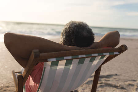Biracial senior man relaxing with hands behind head sitting on folding chair at beach during sunset. lifestyle, love and weekend.の写真素材