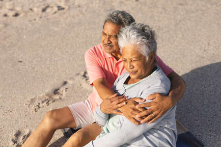Retired senior biracial man embracing woman while sitting together on sand looking away at beach. lifestyle, love and weekend.の写真素材