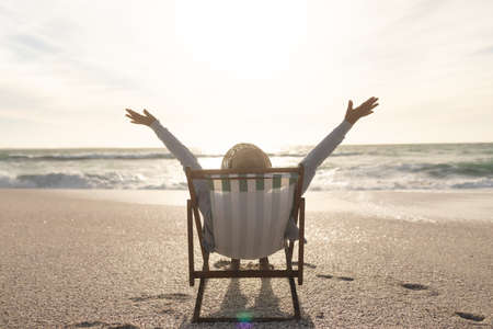 Retired senior biracial woman sitting with hands raised on folding chair enjoying sunset at beach....
