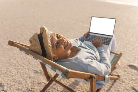 Portrait of smiling senior biracial woman with blank screen laptop showing copy space at beach. lifestyle and telecommunications.の写真素材