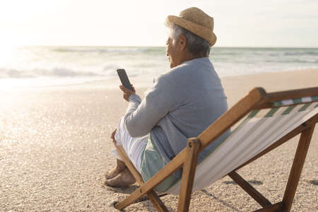 Biracial senior woman using smartphone for video call while sitting on chair at beach during sunset. lifestyle and telecommunications.の写真素材