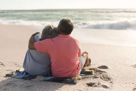 Rear view of senior biracial man sitting with arm around woman enjoying retirement at beach. lifestyle, love and weekend.の写真素材