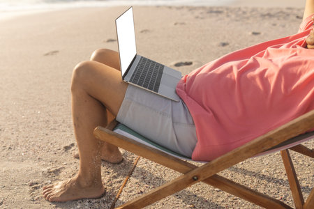 Low section of relaxed senior biracial man sitting with laptop on folding chair at beach. copy space, lifestyle and telecommunications.の写真素材