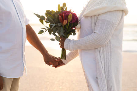 Midsection of multiracial senior couple holding hands with bouquet during wedding ceremony at beach. lifestyle, culture and love.の写真素材