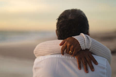 Newlywed biracial senior man and woman hugging each other at beach during sunset. wedding ceremony, lifestyle and love.の写真素材