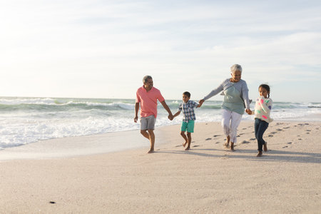 Multiracial grandchildren holding hands with grandparents walking at beach against sky. family,...