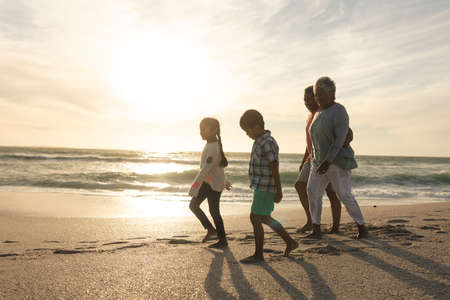 Side view of multiracial grandparents and grandchildren walking on shore at beach during sunset....