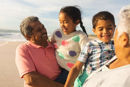 Smiling multiracial grandparents carrying grandchildren enjoying weekend together at sunny beach....