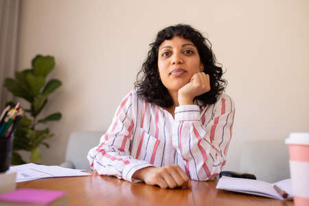 Portrait of young biracial businesswoman leaning with hand on chin at desk in office. female...