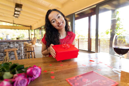 Portrait of happy biracial young woman with gift box at table in restaurant. unaltered, online dating, vitiligo and distant valentine day celebration.の写真素材