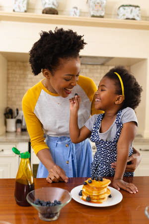 African american daughter feeding berries to her mother in the kitchen at home. family, love and togetherness concept, unaltered.の写真素材