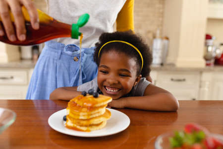 Midsection of mother putting maple syrup on pancake of her smiling african american daughter at home. family, love and togetherness concept, unaltered.の写真素材