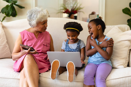 African american grandmother and her two granddaughters using laptop sitting on couch at home. family, love and technology concept, unaltered.の写真素材