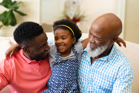 African american grandfather, father and granddaughter smiling looking at each other at home. family, love and togetherness concept, unaltered.の写真素材