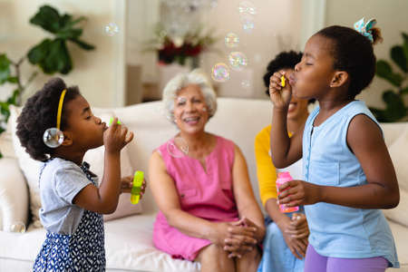 Two african american sisters blowing bubbles while grandmother and mother watching sitting on couch. family, love and togetherness concept, unaltered.の写真素材