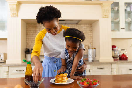 African american mother putting berries over the pancake of her daughter in the kitchen at home. family, love and togetherness concept, unaltered.の写真素材