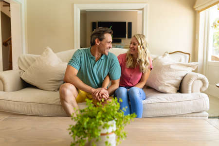Caucasian couple holding hands looking at each other and smiling sitting on the couch at home. couple, love and relationship concept, unaltered.の写真素材