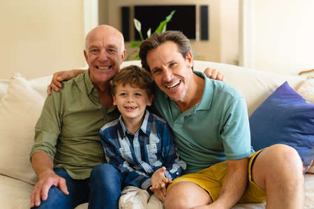 Portrait of caucasian grandfather, father and son smiling while sitting together on couch at home....