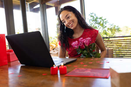 Happy biracial young woman with bouquet video calling through laptop at restaurant. unaltered,...