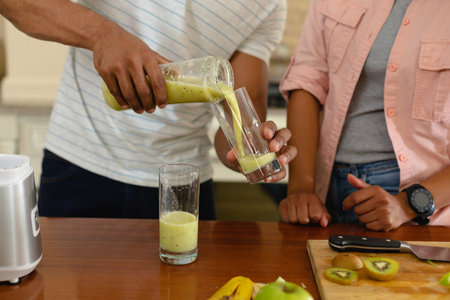 Midsection of young african american man pouring drink in glass from jar at kitchen island. people, love and togetherness concept, unaltered.の写真素材