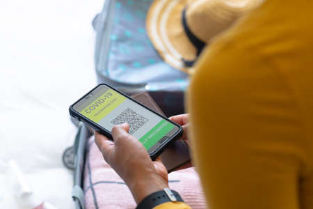 Midsection of african american woman holding covid-19 vaccine certificate on smartphone and passport. people, travel and vaccination concept, unaltered.の写真素材