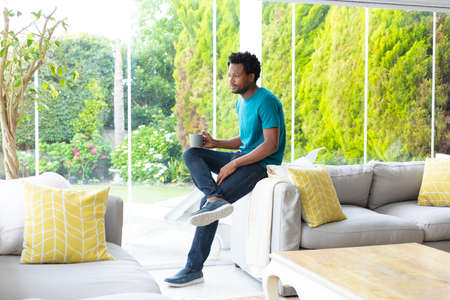 Thoughtful young african american man with coffee mug sitting on sofa in living room at home. people...