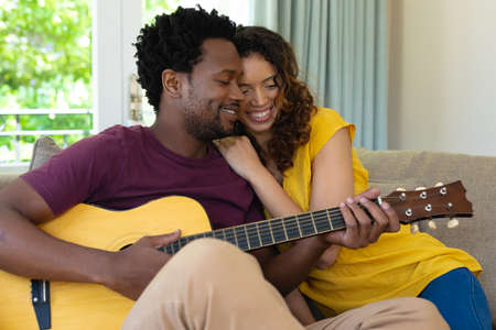 Smiling multiracial couple sitting together with guitar on sofa at home. people, togetherness and music concept, unaltered.の写真素材