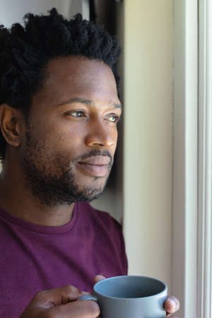 Thoughtful young african american young with coffee mug looking away at home. people and emotions concept, unaltered.の写真素材