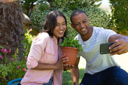 Cheerful young african american couple taking selfie with potted plant through smartphone at garden. people, togetherness and technology, unaltered.の写真素材